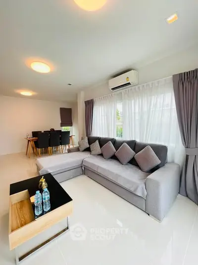 Spacious modern living room with elegant gray sofa and dining area, featuring large windows and natural light.