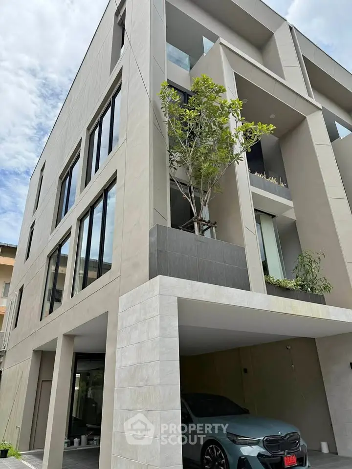 Modern multi-story building with sleek design and carport, featuring large windows and a tree on the balcony.