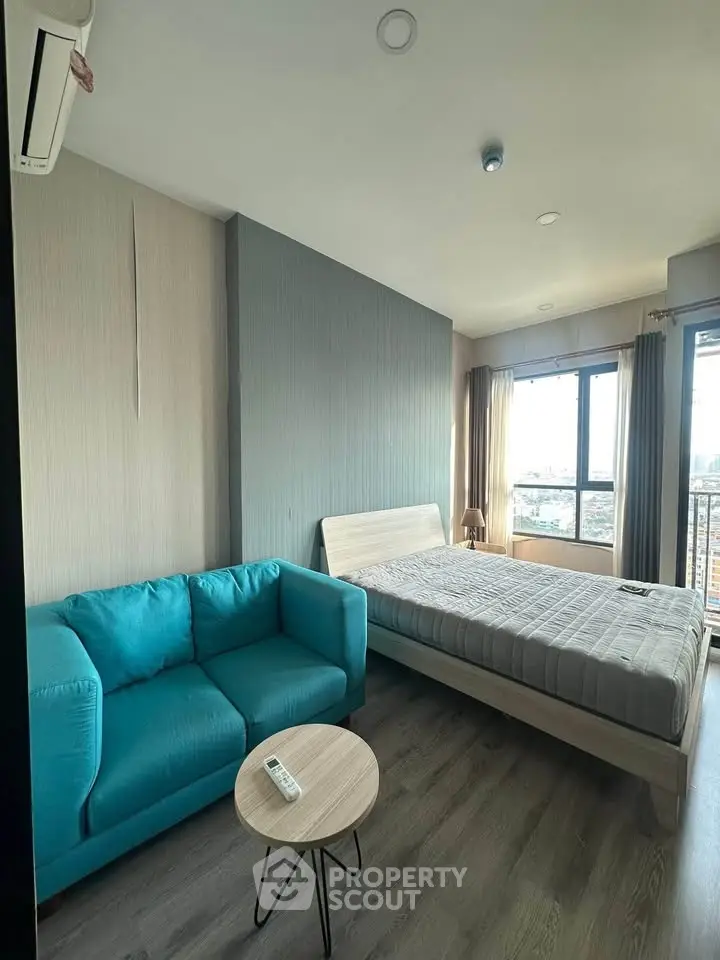 Modern studio apartment with cozy bed and stylish blue sofa, offering a bright city view.