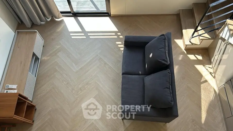 Modern living room with stylish herringbone flooring and cozy gray sofa, featuring natural light and elegant staircase.