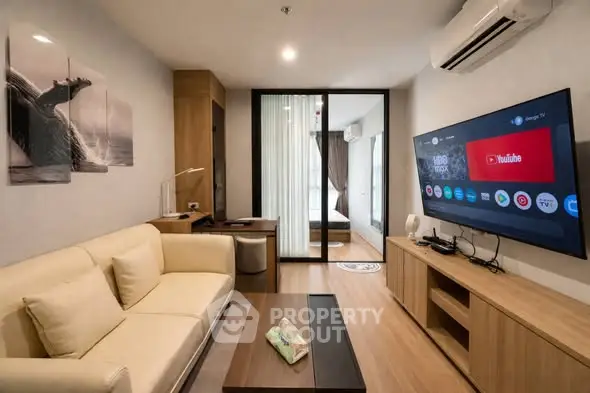 Modern living room with sleek furniture and large TV, open layout leading to bedroom.