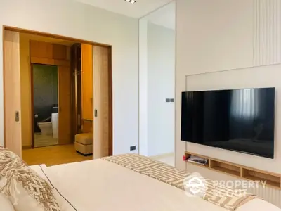 Modern bedroom with sleek design, featuring a large flat-screen TV, wooden accents, and a glimpse of an en-suite bathroom.