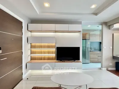 Modern living room with sleek TV unit and open kitchen view, featuring contemporary design and ample lighting.