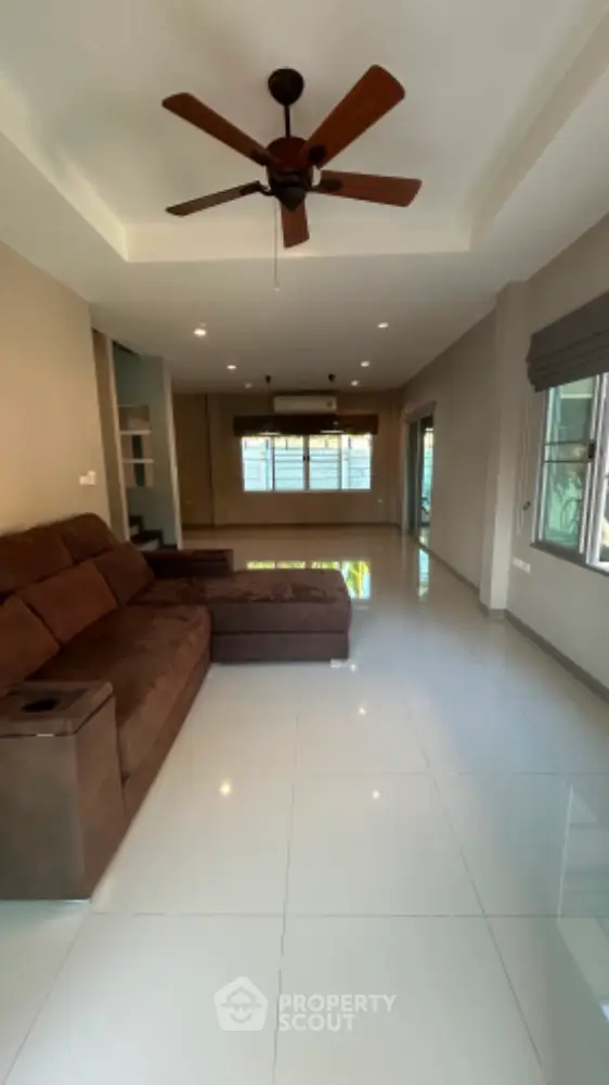 Spacious living room with gleaming white tiles, cozy brown sectional sofa, and a classic ceiling fan, bathed in natural light from large windows.