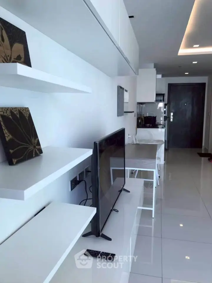 Modern living room with sleek white shelves and flat-screen TV, open layout leading to kitchen area.