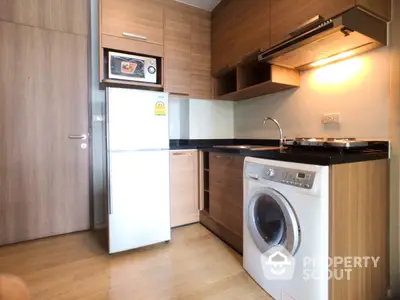 Compact modern kitchen with sleek wooden cabinetry, black countertop, built-in oven, washing machine, and a full-sized refrigerator.