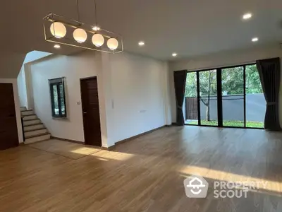 Spacious living room with modern lighting, large windows, and hardwood floors, leading to an inviting staircase and outdoor view.