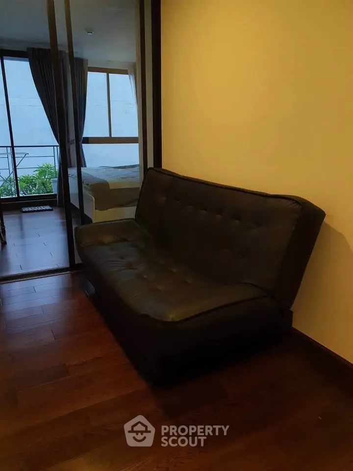 Cozy interior with dark leather sofa and wooden flooring, leading to a bedroom with natural light.