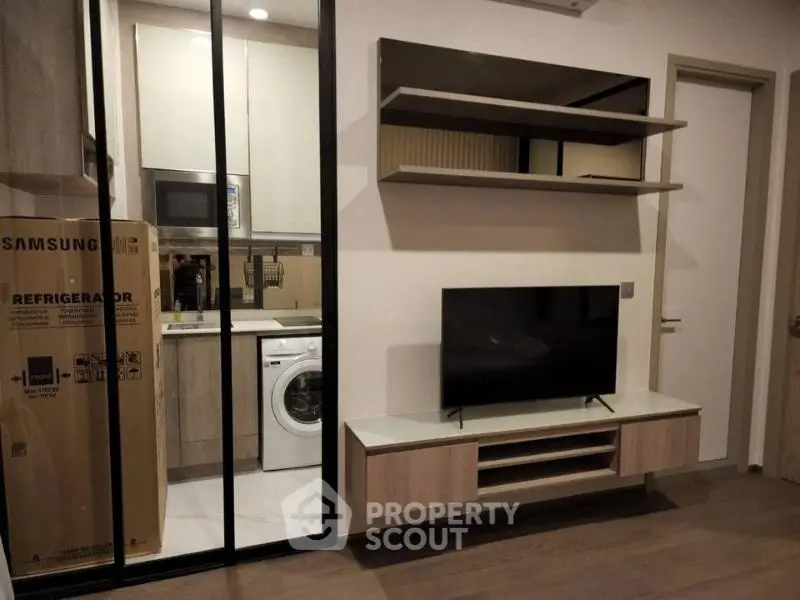 Modern living room with sleek TV unit and view into compact kitchen with appliances.