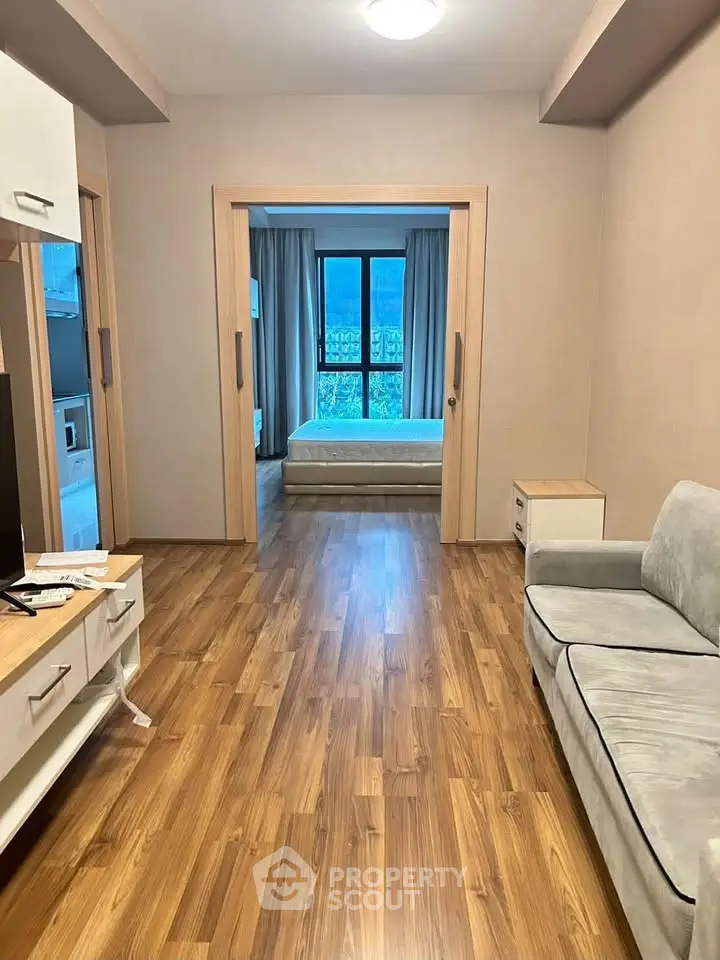 Spacious living room with wooden flooring leading to a cozy bedroom with large windows.