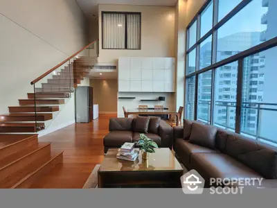 Luxurious double-height living room with modern leather sofas, expansive windows offering city views, and elegant wooden staircase leading to an upper level.