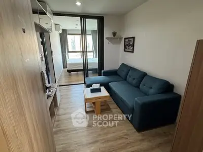 Modern living room with cozy blue sofa and wooden flooring, leading to a bedroom with glass partition.
