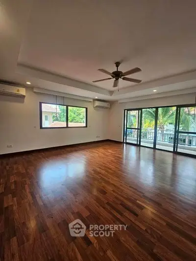 Spacious living room with wooden flooring and large windows opening to a balcony.