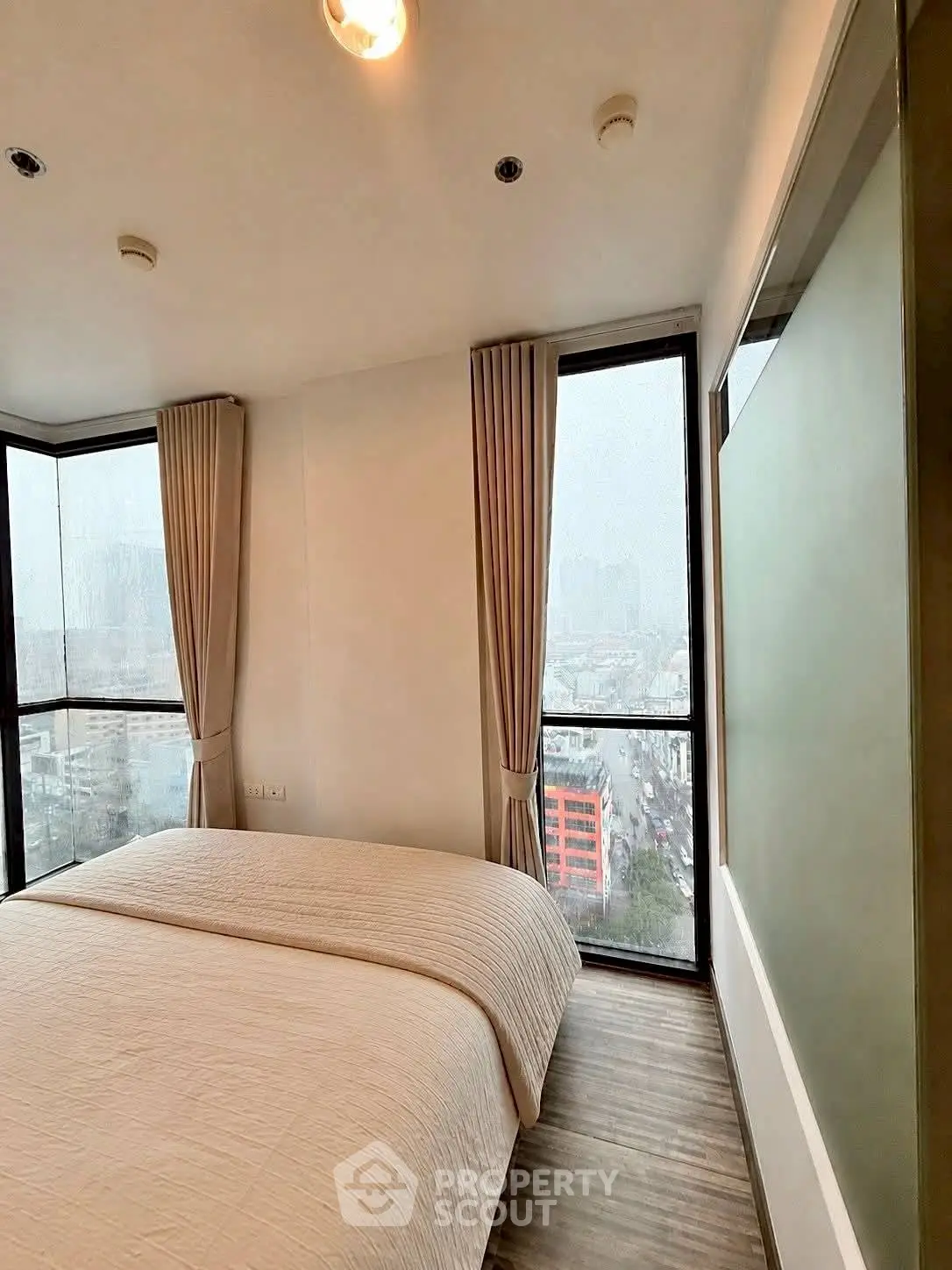 Modern bedroom with large windows offering city views, featuring elegant curtains and cozy bedding.