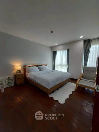 Cozy bedroom with wooden flooring, modern furniture, and natural light from large windows.
