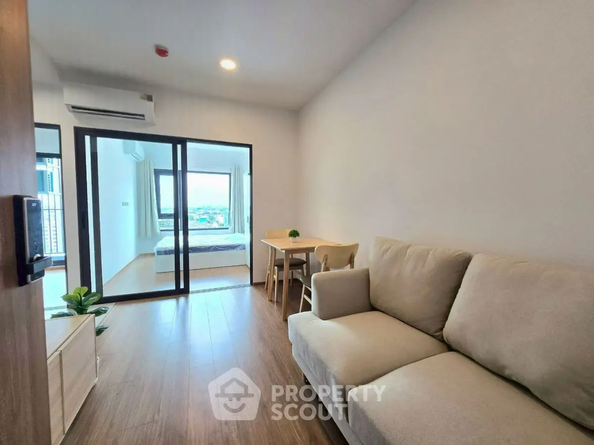 Modern apartment living room with cozy sofa and wooden flooring, leading to a bright bedroom with a view.
