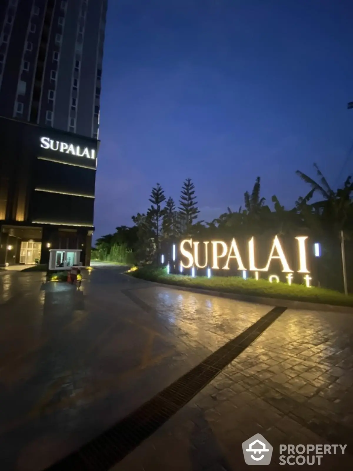 Modern high-rise building entrance at dusk with illuminated signage and lush greenery.