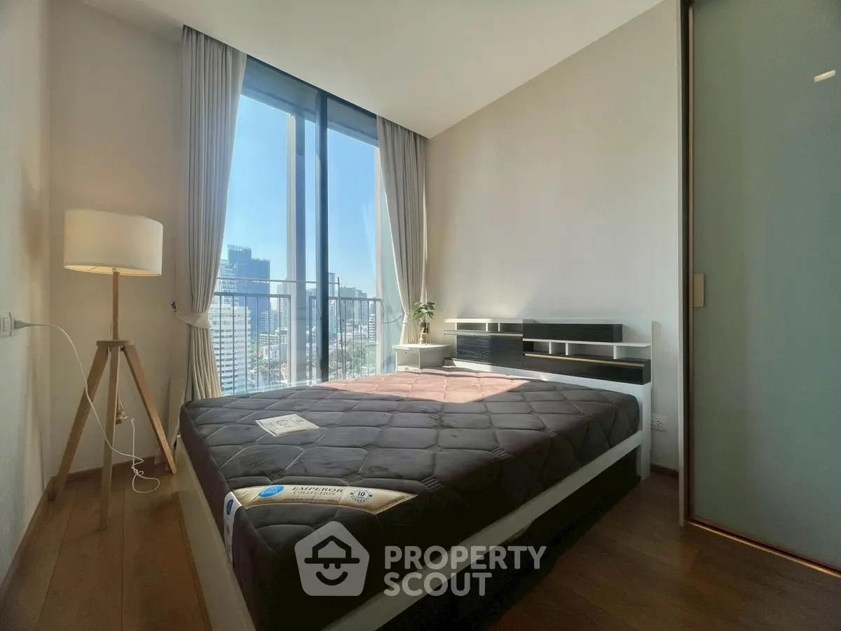 Modern bedroom with large window offering city view, featuring sleek furniture and natural light.