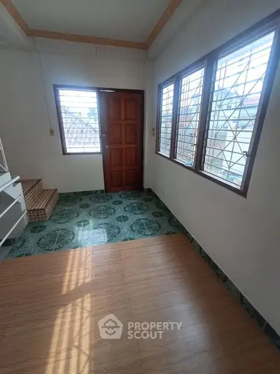 Spacious room with large windows and wooden door, featuring green patterned flooring and natural light.