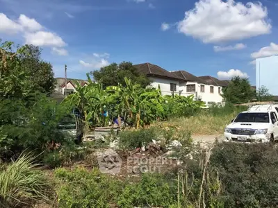 Spacious residential land with lush greenery and nearby houses under a clear blue sky.