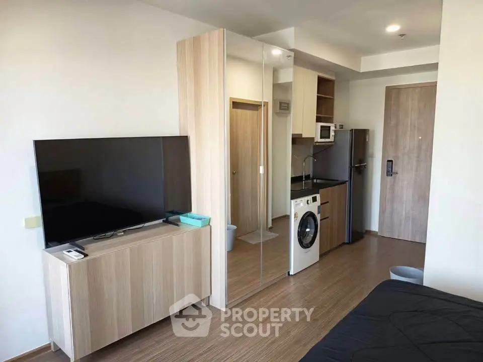 Modern studio apartment with sleek furniture, TV, and compact kitchen featuring washing machine and microwave.