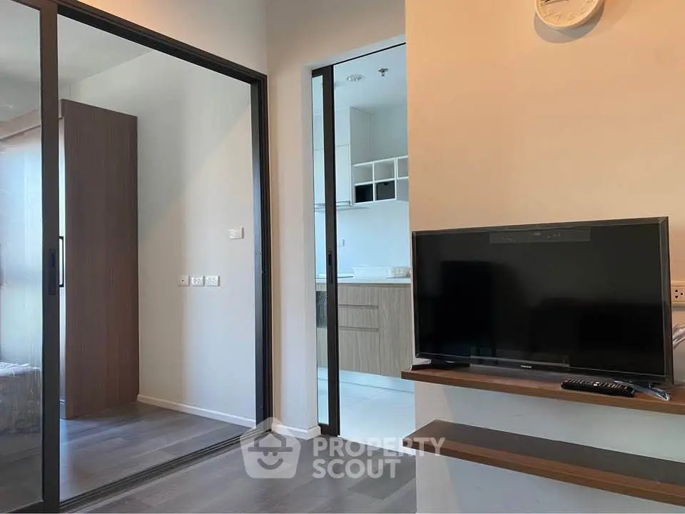 Modern apartment interior with sleek TV setup and glass sliding doors leading to a bright kitchen area.