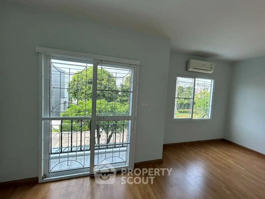 Bright empty room with large windows and wooden flooring, overlooking lush greenery.