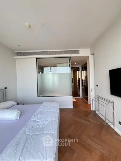 Spacious modern bedroom with wooden flooring and large window, featuring minimalist decor and a wall-mounted TV.