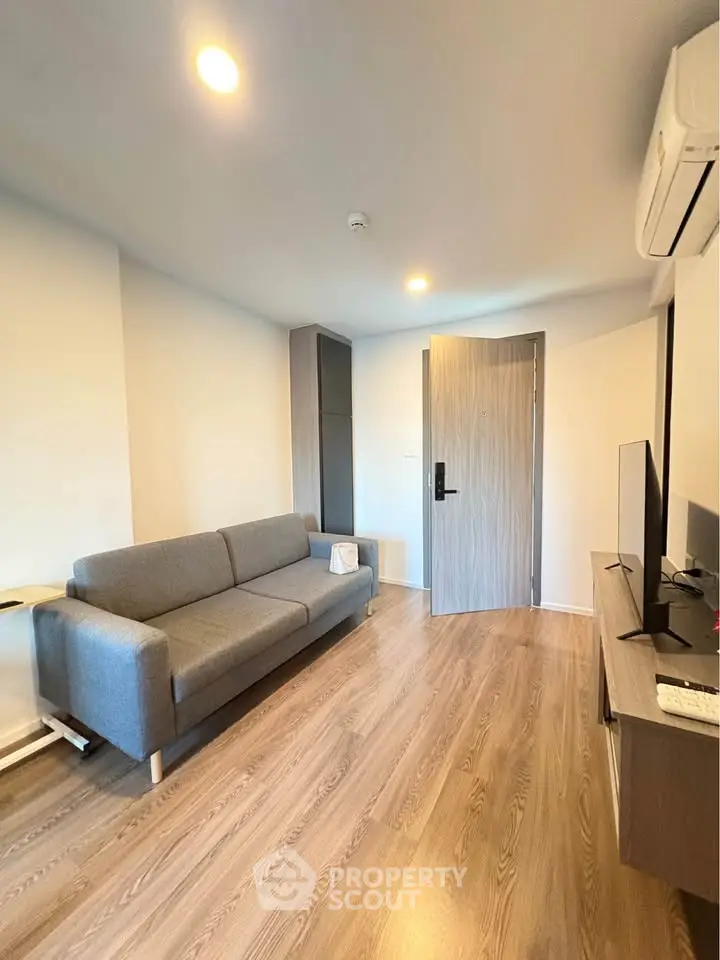 Modern living room with sleek gray sofa and wooden flooring, featuring a flat-screen TV and air conditioning.