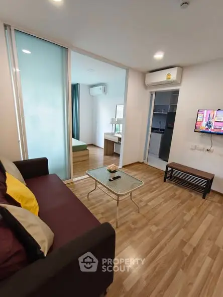 Modern living room with wooden flooring, glass coffee table, and view into bedroom and kitchen.