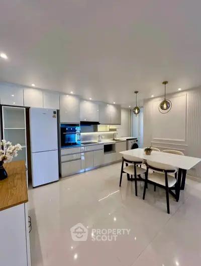 Modern kitchen with sleek white cabinets and dining area, featuring elegant lighting.