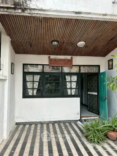 Charming entrance with wooden ceiling and tiled pathway, inviting green door, and lush plants.