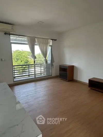 Spacious living room with large window and wooden flooring, offering a serene view of greenery.