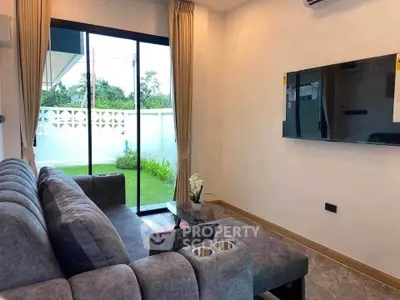 Modern living room with plush gray sofa and wall-mounted TV, overlooking a lush green garden through sliding glass doors.