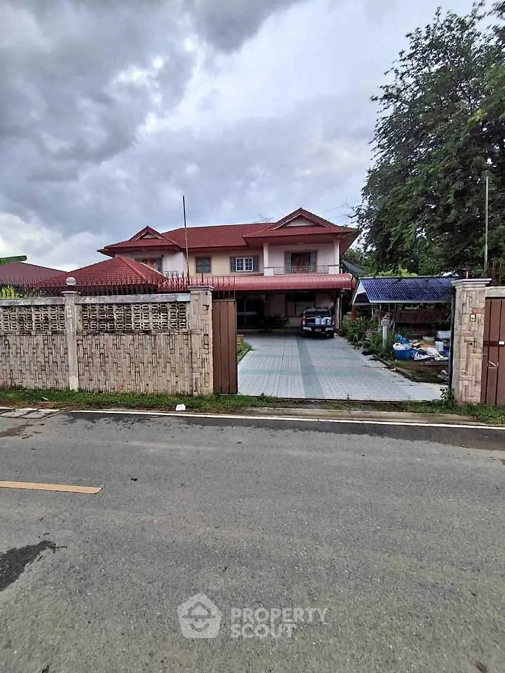 Charming two-story house with red roof and spacious driveway, surrounded by lush greenery and secure fencing.