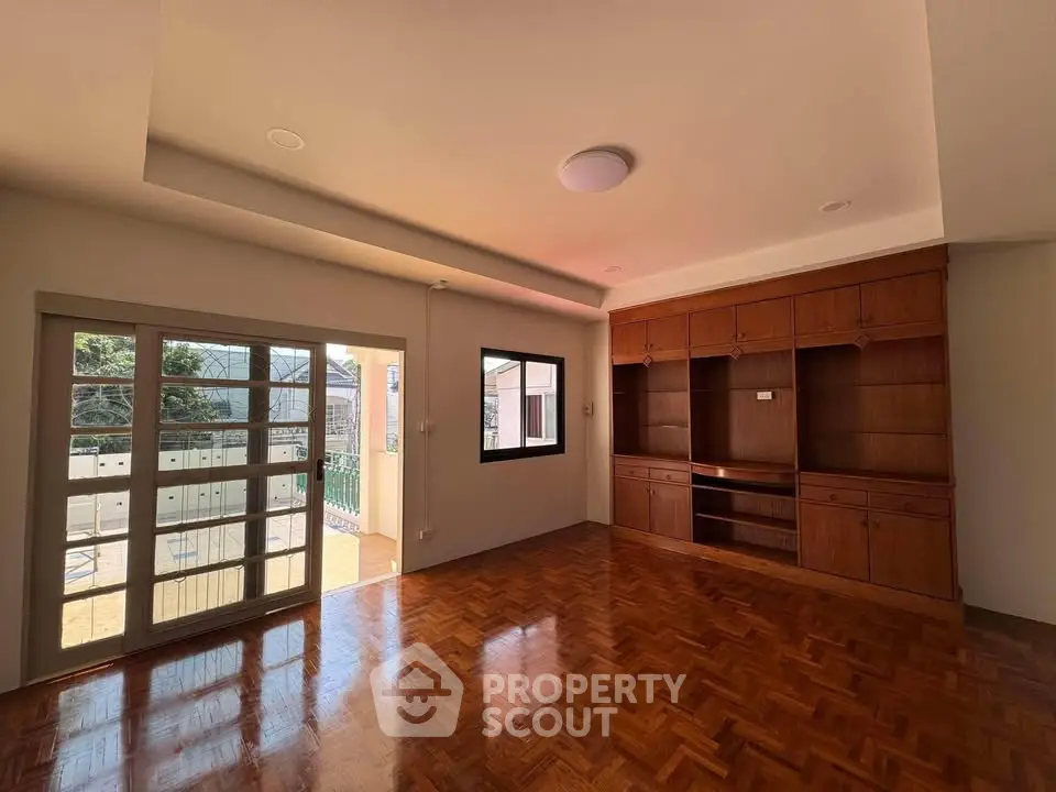 Spacious living room with wooden flooring and built-in shelves, opening to a sunny balcony.