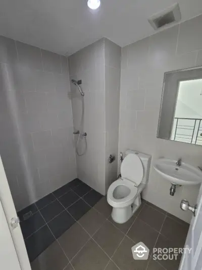 Modern bathroom with sleek gray tiles, wall-mounted shower, classic white toilet, and pedestal sink, featuring a small window for natural light.