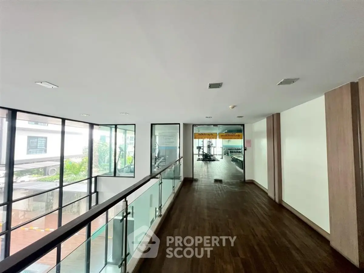 Spacious modern corridor leading to a well-equipped gym with glass walls and wooden flooring.