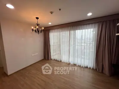 Spacious and well-lit living room with elegant hardwood flooring and large windows draped with sheer curtains and luxurious dark curtains, complemented by a classic chandelier.