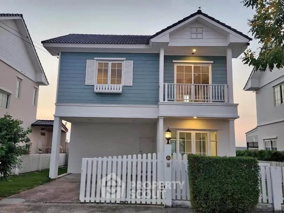 Charming two-story house with blue siding and white picket fence, featuring a cozy balcony and warm exterior lighting.