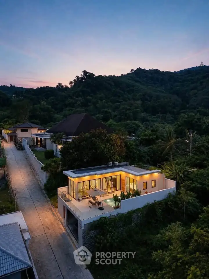 Stunning modern villa with panoramic mountain views at sunset, featuring expansive glass windows and lush surroundings.