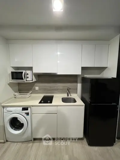 Modern compact kitchen with sleek white cabinets, washing machine, microwave, and black fridge.
