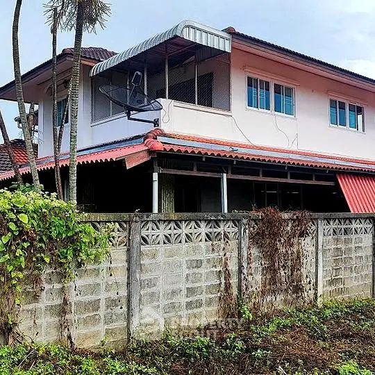 Charming two-story home with a spacious balcony and satellite dish, surrounded by lush greenery and a sturdy concrete fence for privacy.