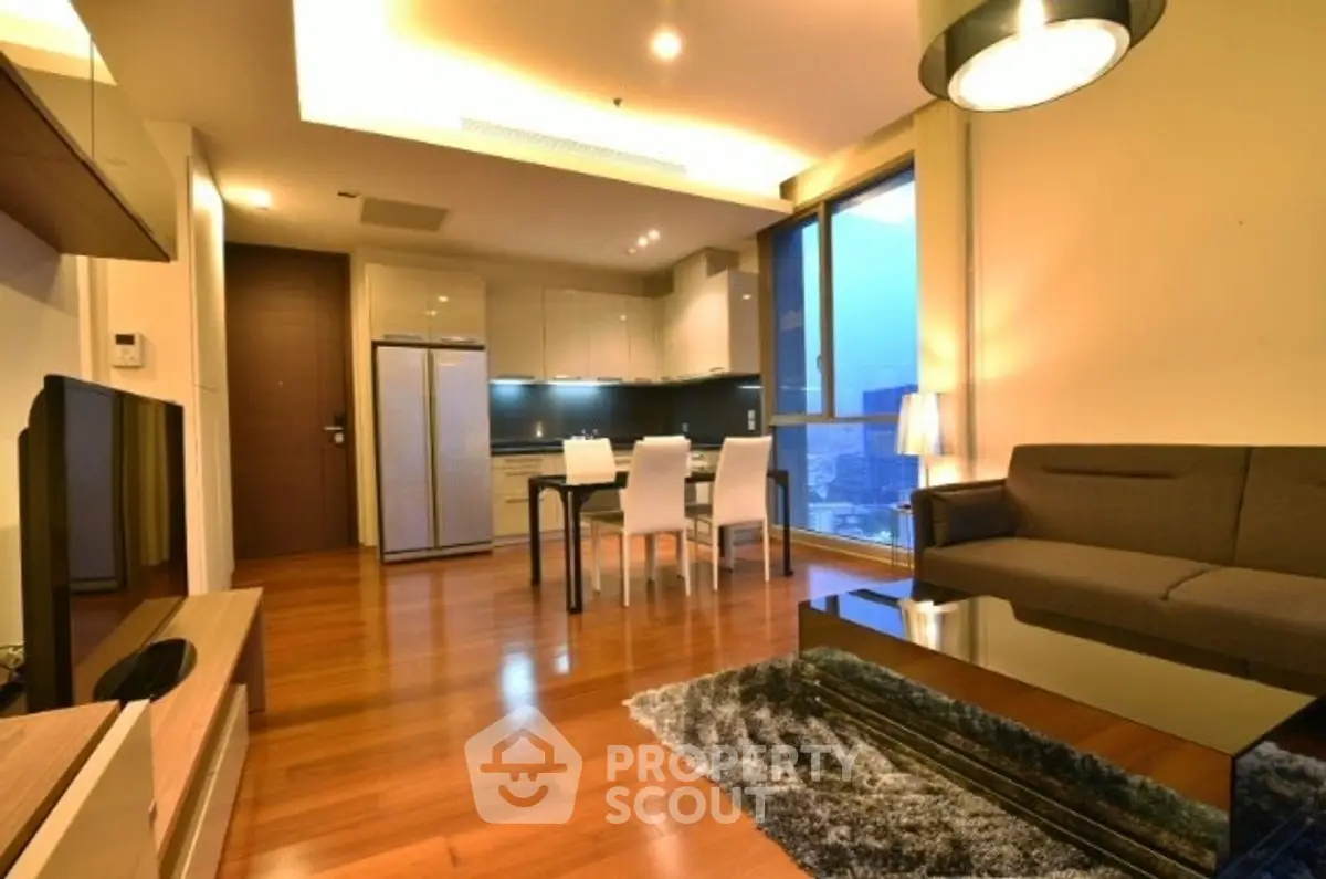 Modern living room with open kitchen and city view, featuring sleek furniture and warm lighting.