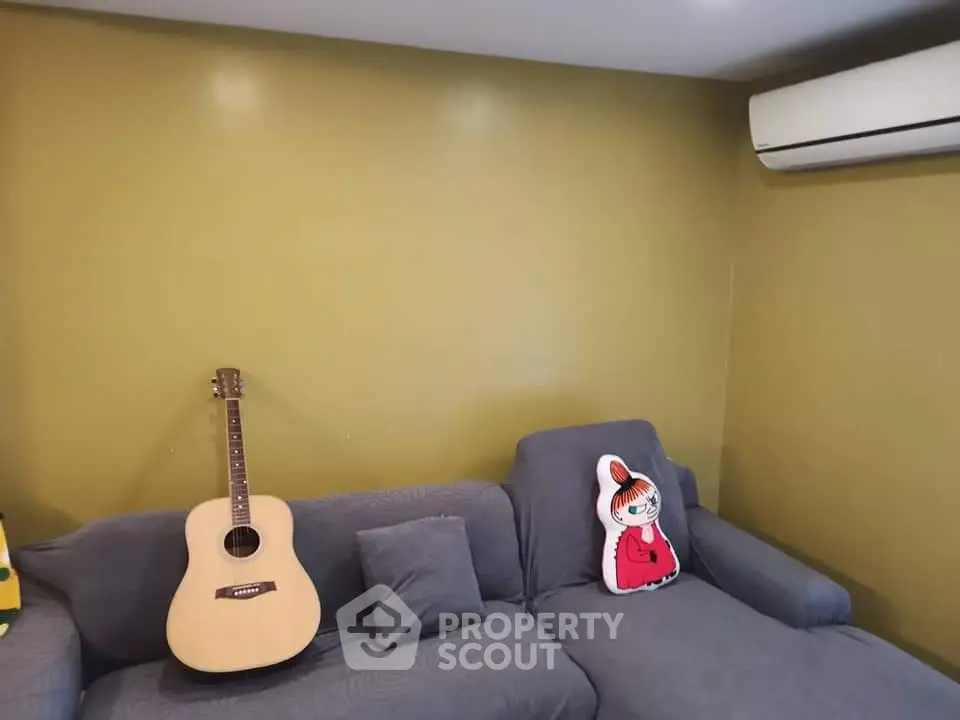 Cozy living room with gray sofa, guitar, and air conditioning unit on mustard wall.