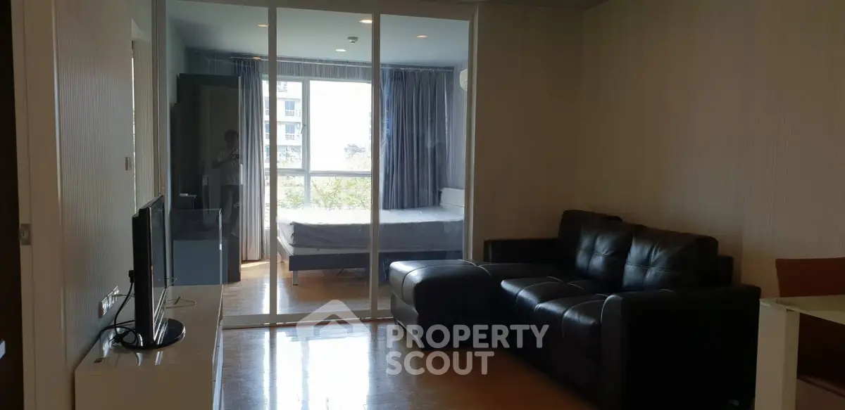 Modern living room with glass partition leading to a cozy bedroom, featuring a sleek black leather sofa and natural light.