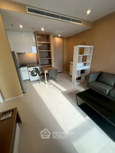 Modern living room with compact kitchen and washing machine, featuring sleek furniture and ample natural light.