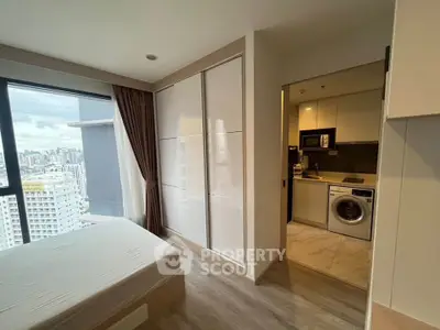 Modern apartment bedroom with city view and adjacent kitchen featuring washing machine and microwave.