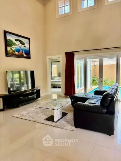 Spacious living room with high ceilings and modern decor, featuring large windows and a sleek black leather sofa.