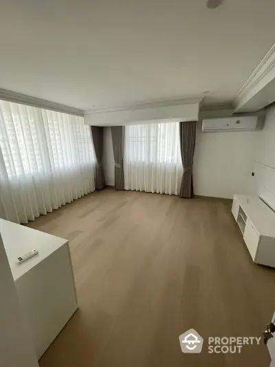 Spacious and well-lit living room with large windows draped in elegant curtains, featuring a modern air conditioning unit and sleek white cabinetry, perfect for comfortable urban living.